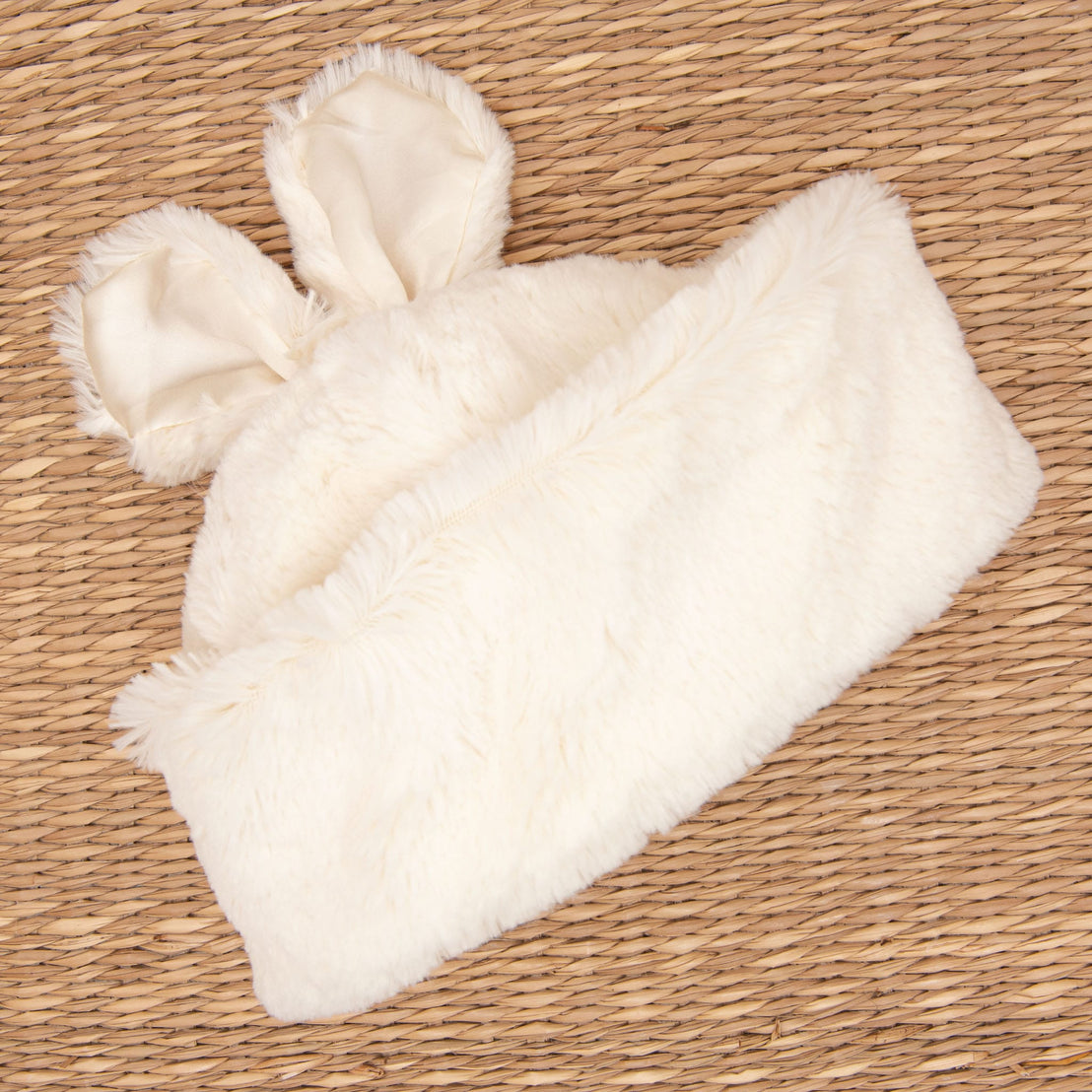A Glad Dreams Coat Set, featuring a white, furry hat with bunny ears, rests on a woven straw mat background. The plush texture of the set exudes warmth and coziness, making it a unique baby gift for chilly days.