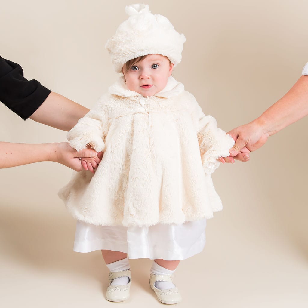 A baby is dressed in the luxurious Glad Dreams Coat Set, featuring a cream-colored faux fur coat and matching hat, and supported by two adults. The ensemble is perfectly completed with white shoes and socks. The little one gazes directly at the camera, embodying the essence of a unique baby gift for winter warmth.