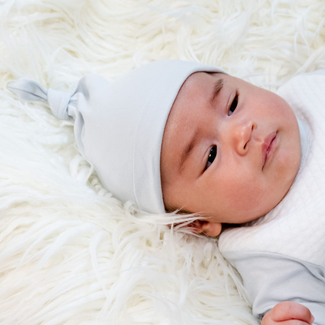 A baby lies on a fluffy white blanket, adorned in the Grayson Newborn Gift Set, which includes a soft pima cotton gown. With a light gray knotted hat completing the matching ensemble, the baby gazes to the side with a calm expression, creating an ideal scene for a personalized baby gift. Plus, you can save 10% on this beautiful set.