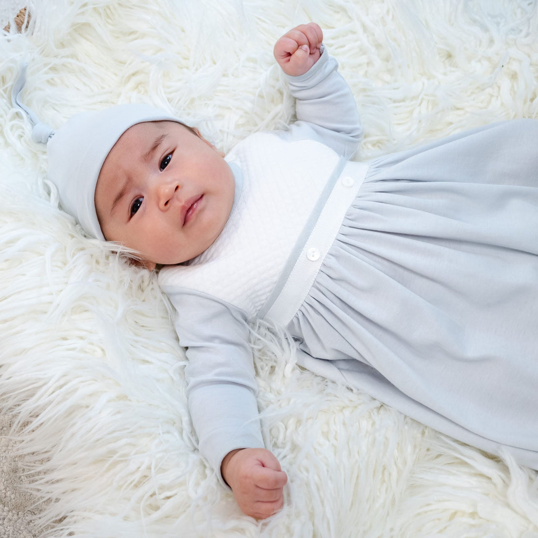 A baby dressed in a light blue Grayson Newborn Gift Set - Save 10%, made from soft pima cotton, along with a matching hat, is lying on a plush white textured surface. With a calm expression and one arm slightly raised, the baby gazes upward, epitomizing the ideal personalized baby gift.