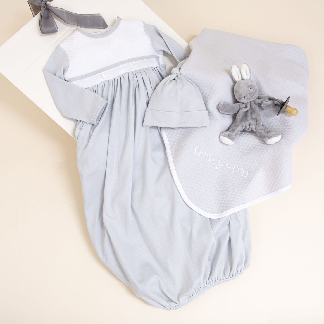 The Grayson Newborn Gift Set, now available with a 10% discount, includes a light gray pima cotton gown with long sleeves and a matching hat displayed on a cream surface. Completing the personalized baby gift set is an embroidered gray blanket with "Grayson" and a plush rabbit toy equipped with a pacifier holder, all set in an elegantly subtle nursery environment.