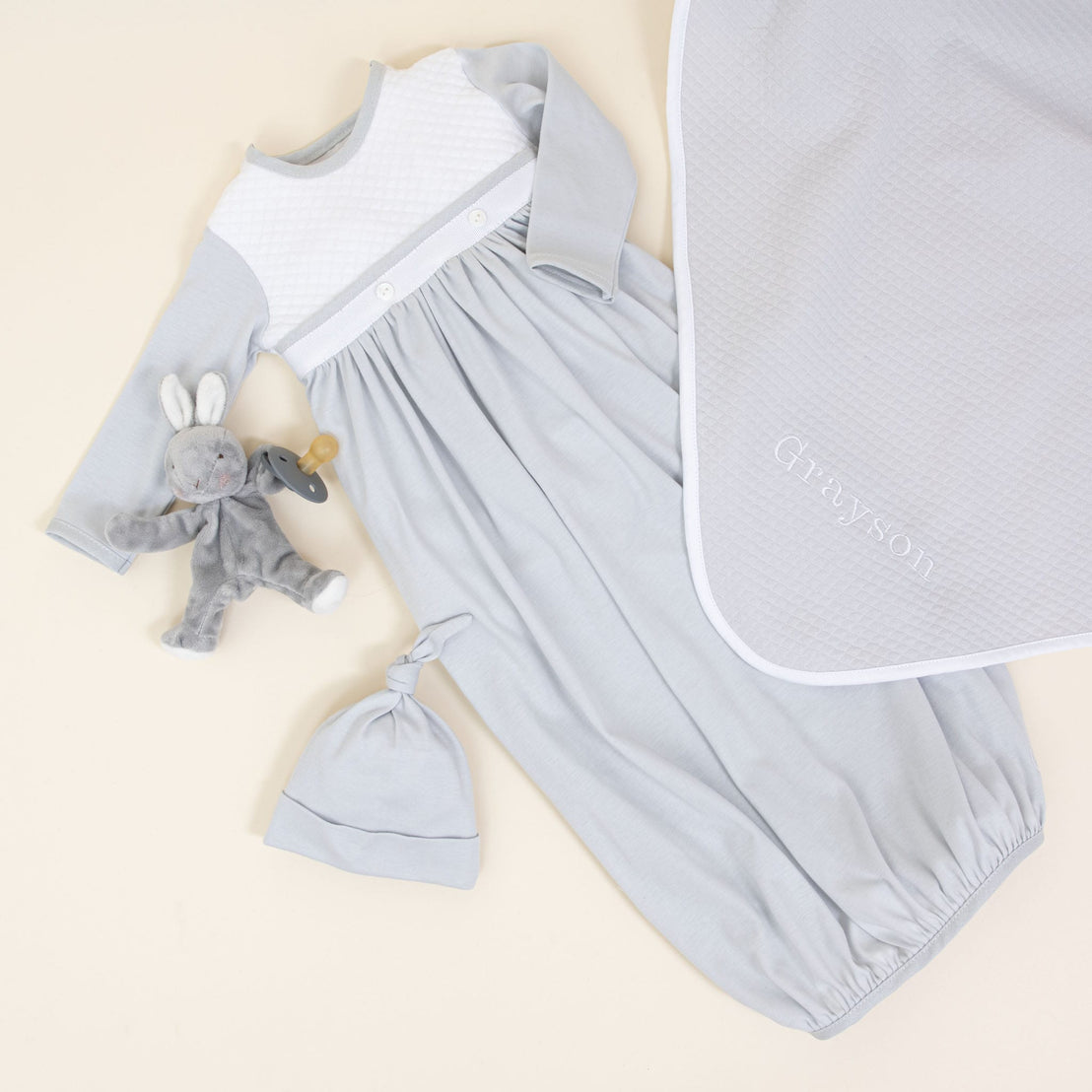 The Grayson Newborn Gift Set - Save 10% features a light blue newborn gown crafted from soft pima cotton, complete with long sleeves and white detailing, elegantly displayed on a surface. Accompanying the gown is a matching baby hat, perfectly enhancing the arrangement along with a gray plush bunny toy equipped with a pacifier and an embroidered gray blanket, making it an ideal personalized baby gift.