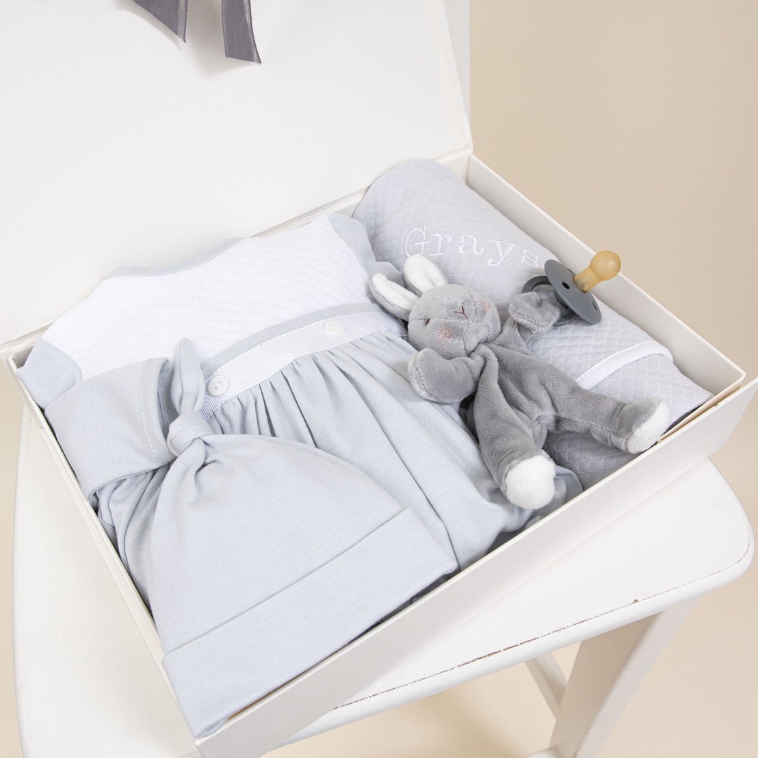 Displayed on a white chair is the Grayson Newborn Gift Set—now available at 10% off—which includes a soft pima cotton newborn gown, matching hat, little plush toy rabbit, and a pacifier with holder, all personalized with the name "Grayson" and presented in an elegant white gift box.