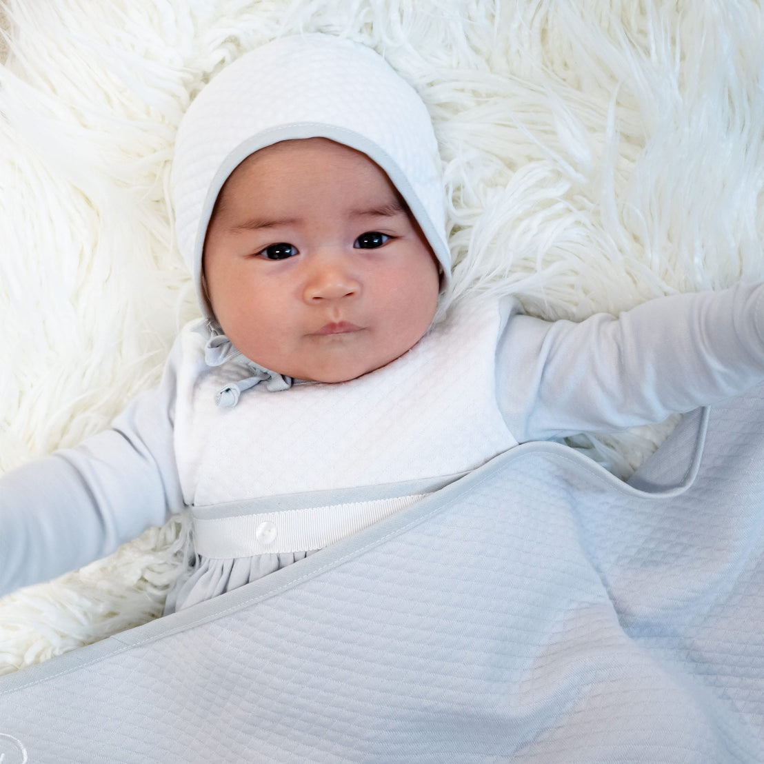 A baby in a quilted cotton ensemble, featuring the Grayson White Quilted Bonnet, lies peacefully on a fluffy white surface with outstretched arms and a calm expression.