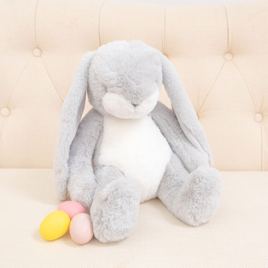 A fluffy Grey Nibble Bunny with a white belly sits on a cream-colored sofa. Its embroidered face beams next to three pastel-colored eggs: yellow, pink, and orange. Made with Baby Safe Soft Plush, it's perfect for snuggles.