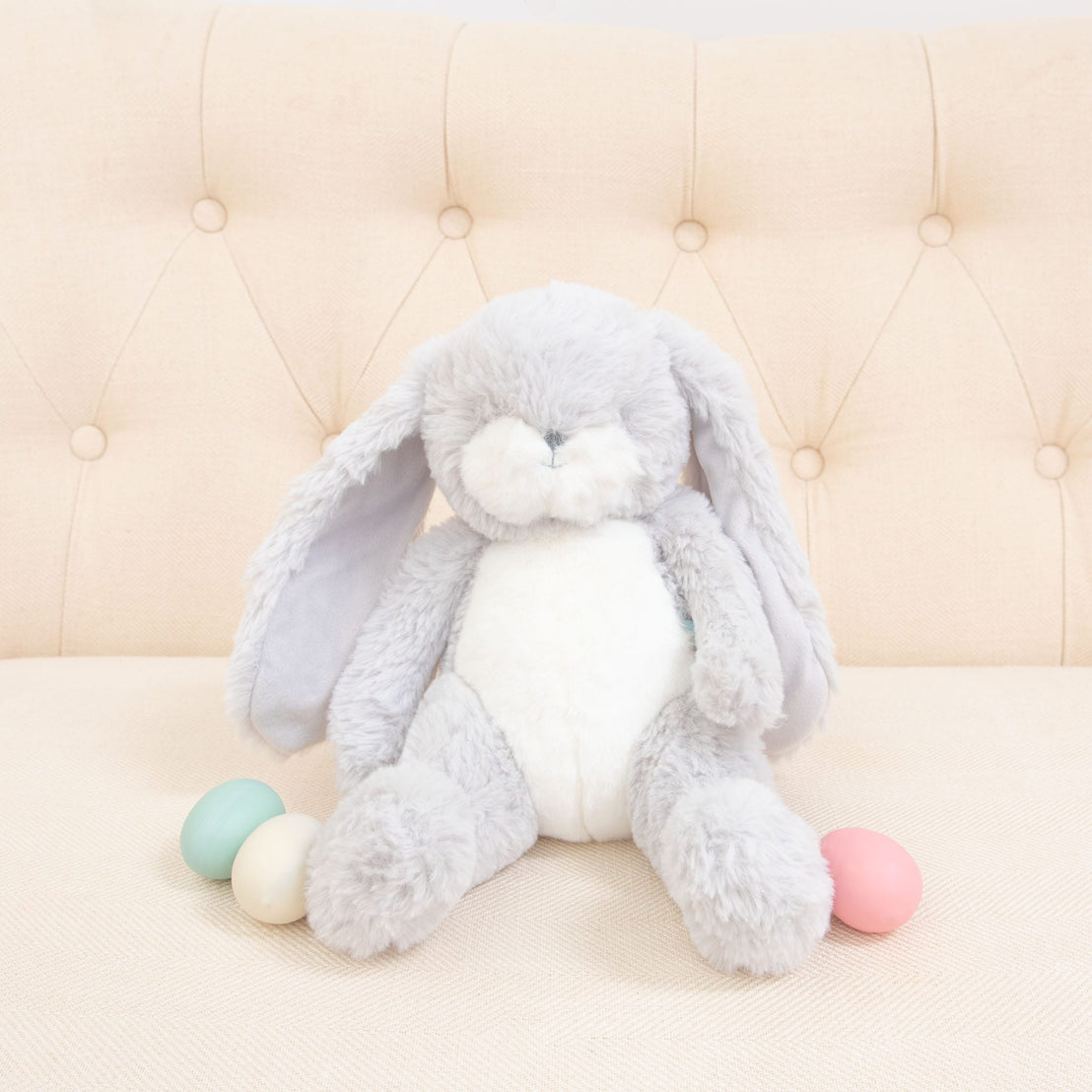 The Grey Nibble Bunny, featuring an embroidered face and floppy ears, sits on a cream-colored tufted couch. Positioned next to it are three pastel-colored eggs in shades of blue, mint, and pink, creating a delightful springtime scene with this baby-safe soft plush.