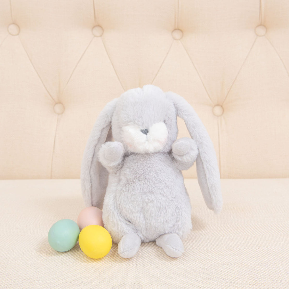 A fluffy Grey Nibble Bunny with an embroidered face and long ears sits on a beige tufted cushion. Next to it are four pastel-colored eggs: pink, green, blue, and yellow. The soft and neutral background enhances the cozy atmosphere of these baby-safe soft plush companions.