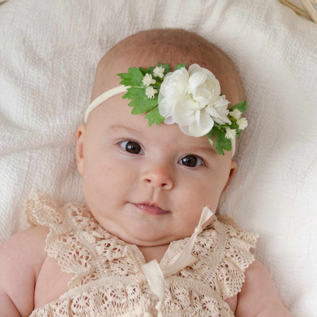 A baby in a cream lace dress and the Guinevere Flower Headband, featuring delicate handmade ivory flowers, lies on a soft, white blanket. The baby gazes gently towards the camera.