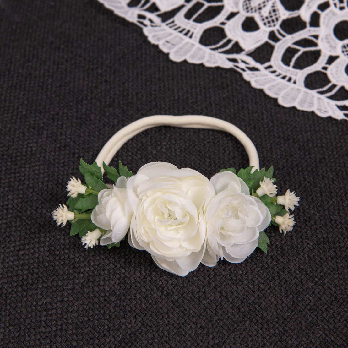 The Guinevere Flower Headband, adorned with ivory flowers and green leaves, rests on a dark fabric surface. Nearby, a piece of white lace subtly enhances the delicate ambiance.