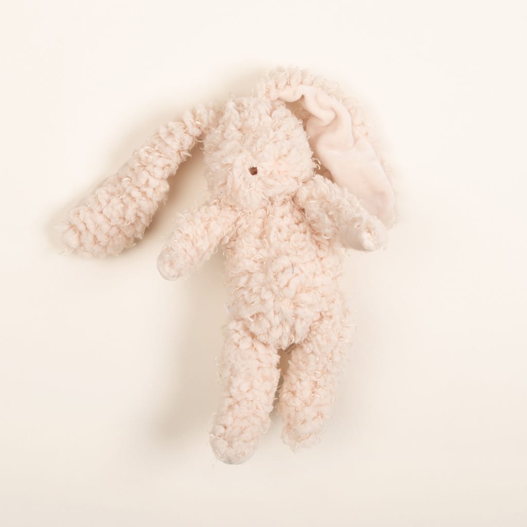 Harey Bunny, a plush toy with curly pale pink fur and floppy ears, rests on a light cream background. It has an embroidered face, with one ear bent over and the other sticking out to the side. This minimalist soft bunny is an ideal gift for babies.