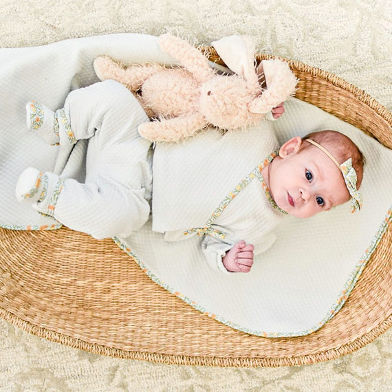 A baby lies in a woven basket on a cream-colored blanket, dressed in a light gray outfit with floral trim and a matching headband. The baby is holding the Harey Bunny, which features an embroidered face. The basket rests on a textured surface, creating the ideal setting for a baby gift.