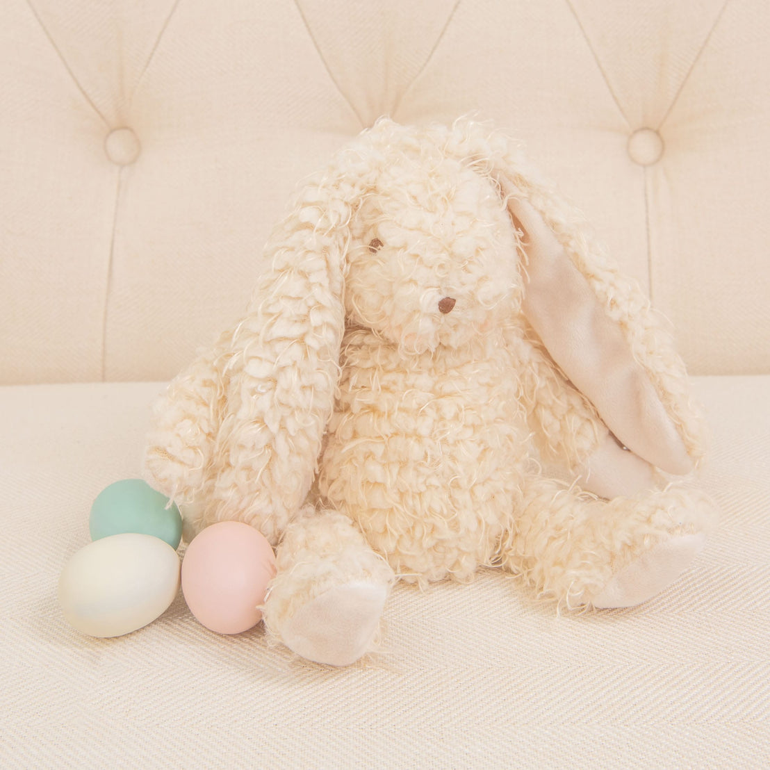 A plush Harey Bunny, featuring cream-colored and soft almond scraggle fur, is sitting on a light-colored sofa. With its long ears, it's joined by three pastel-colored eggs—green, pink, and off-white. This delightful arrangement serves as an ideal baby gift against the tufted upholstery backdrop.