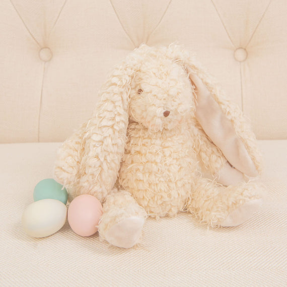 A plush Harey Bunny, featuring cream-colored and soft almond scraggle fur, is sitting on a light-colored sofa. With its long ears, it's joined by three pastel-colored eggs—green, pink, and off-white. This delightful arrangement serves as an ideal baby gift against the tufted upholstery backdrop.