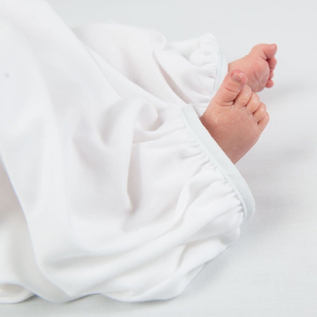 A close-up of tiny newborn feet peeking out from the soft, gathered hem of the Harrison Newborn Gown, made from textured white fabric.