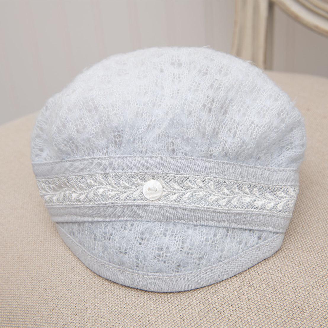 Harrison Newborn Knit Hat displayed on a neutral-toned cushion, showcasing its soft, textured knit in blue with lace trim and a button accent.