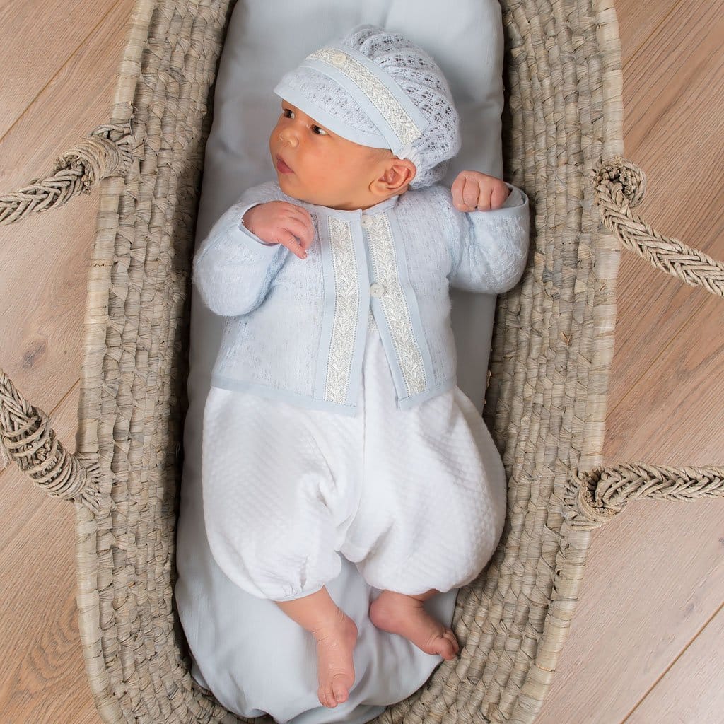 A baby lying in a woven bassinet, dressed in the Harrison Newborn Sweater paired with a matching knit hat and quilted romper.