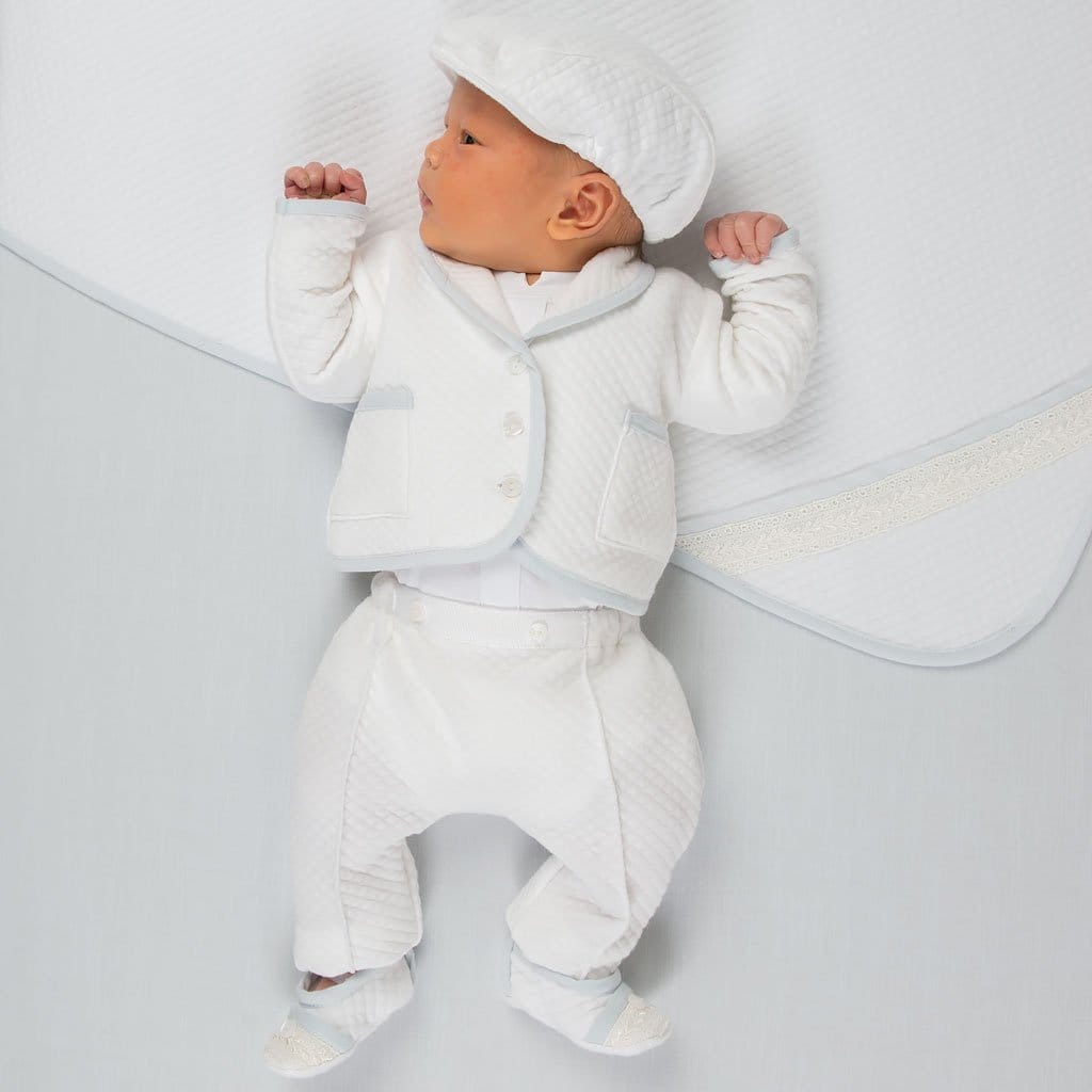 A baby dressed in the Harrison Quilted Newborn Suit, featuring a textured white quilted jacket with blue trim, matching pants, and a coordinating cap, lying on a matching quilted blanket.