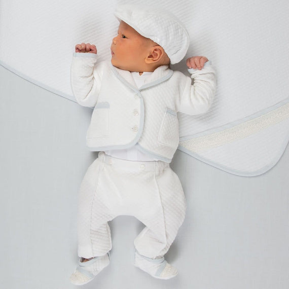A baby dressed in the Harrison Quilted Newborn Suit, featuring a textured white quilted jacket with blue trim, matching pants, and a coordinating cap, lying on a matching quilted blanket.