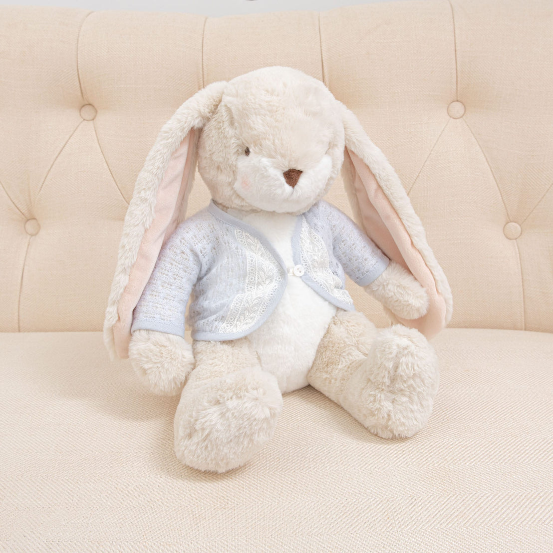 The Harrison Sweater Bunny, a plush toy with long ears and soft, cream-colored fur, dressed in a light blue sweater sits on a tufted beige couch.