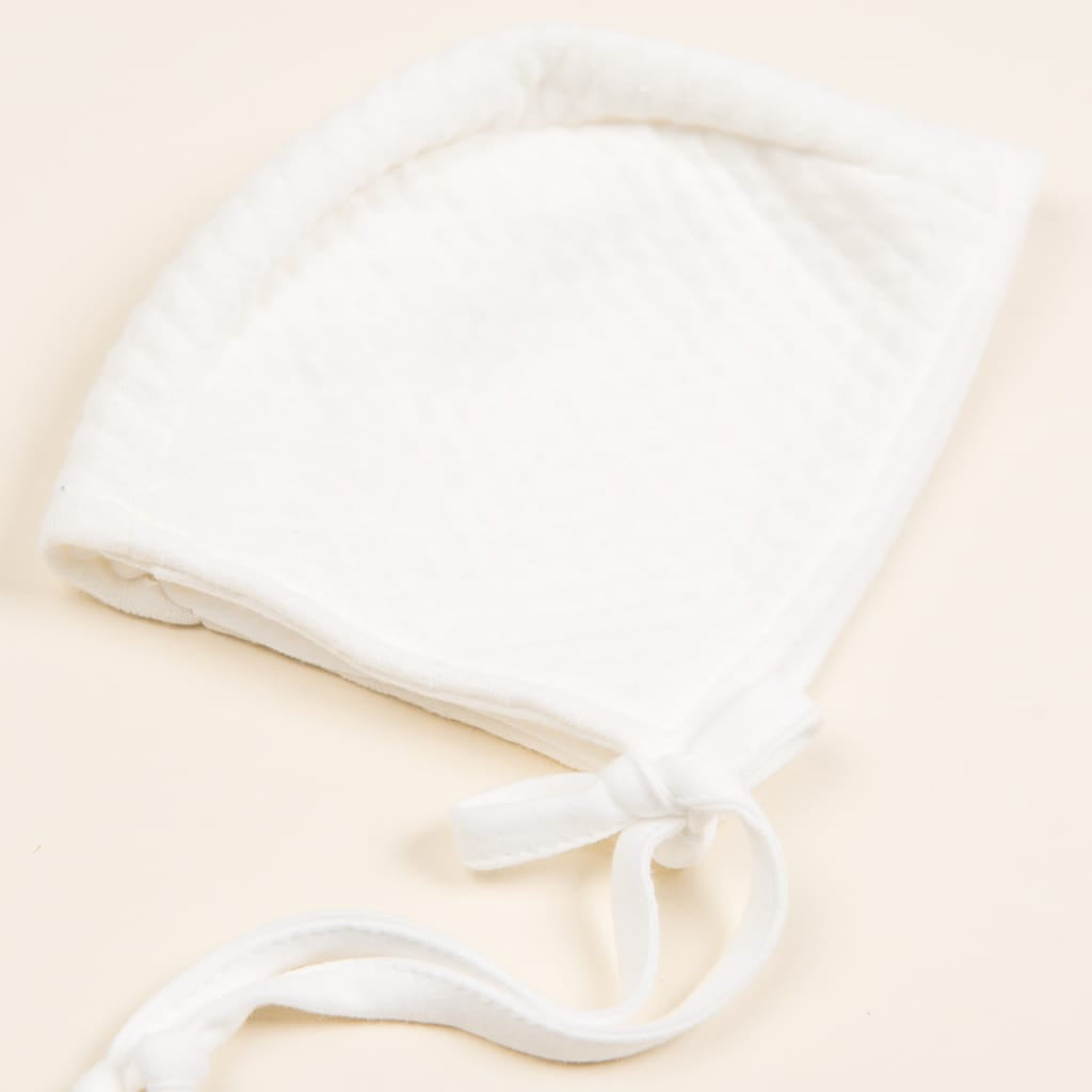 The Ivory Quilt Bonnet, crafted from luxurious Pima cotton, features a soft quilted texture with a tie strap and rests gracefully on a light cream background. Its rounded shape with gently curved edges makes it perfect for any newborn outfit.