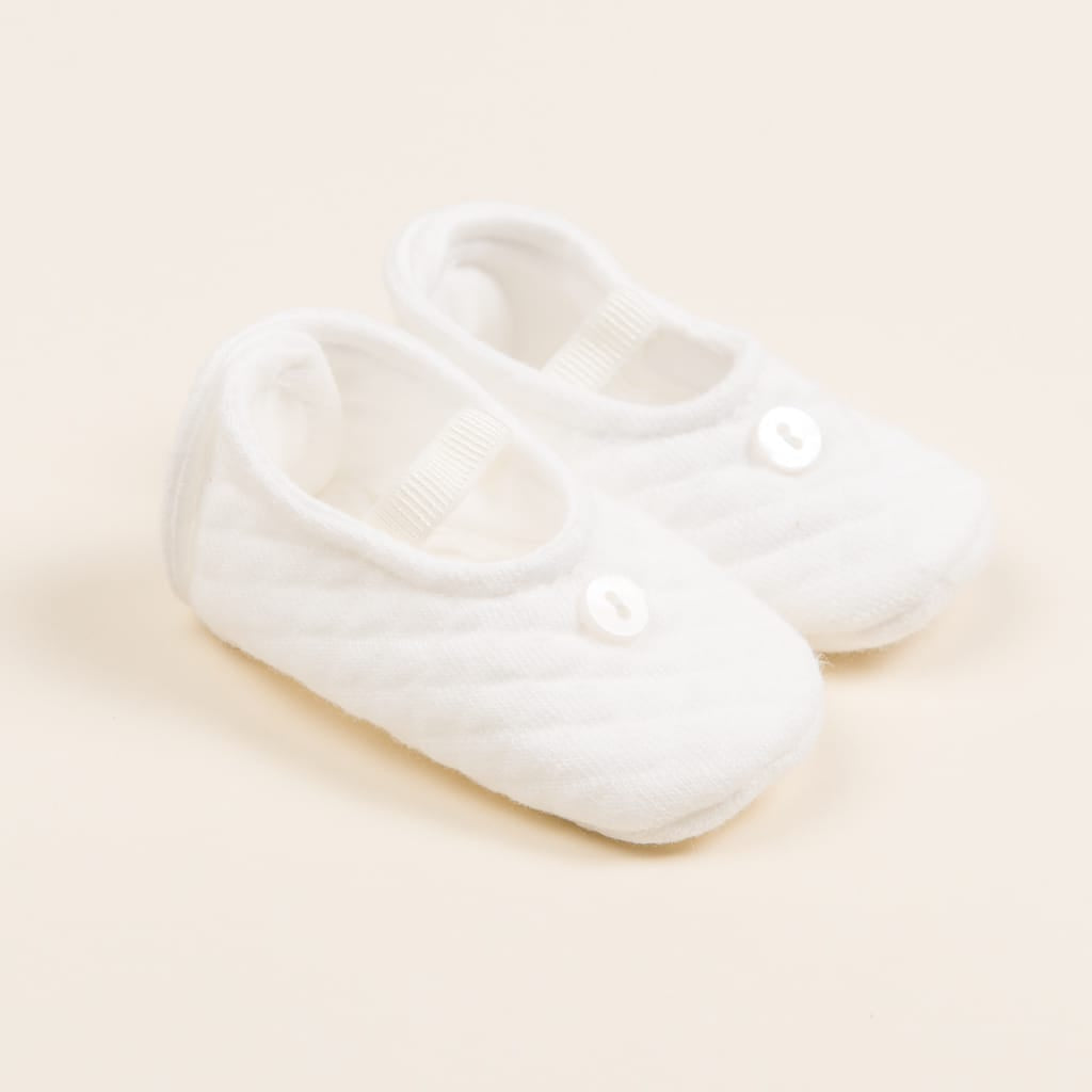 A pair of soft Ivory Quilted Booties crafted from textured cotton, featuring a small white button on each shoe. These handmade baby shoes include elastic straps over the top, all set against a light beige background.