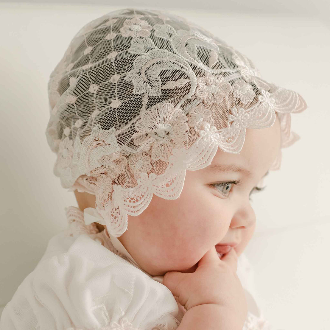 Baby wearing a Joli Sheer Bonnet and matching dress looks away from the camera with one hand in her mouth. The side profile showcases the intricate details and embroidered netting. 