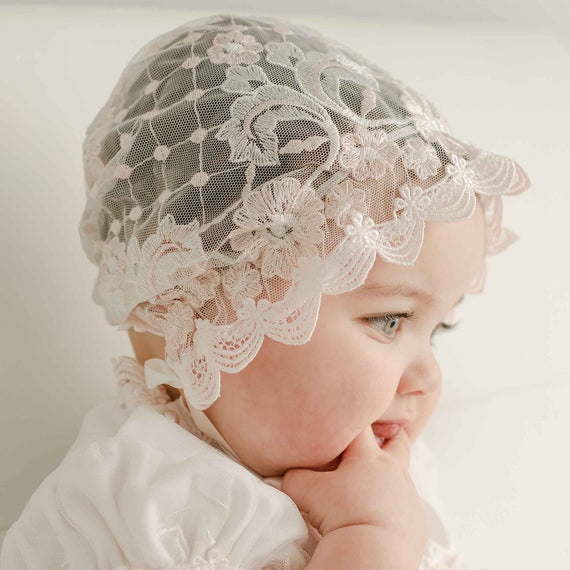 Baby wearing a Joli Sheer Bonnet and matching dress looks away from the camera with one hand in her mouth. The side profile showcases the intricate details and embroidered netting. 