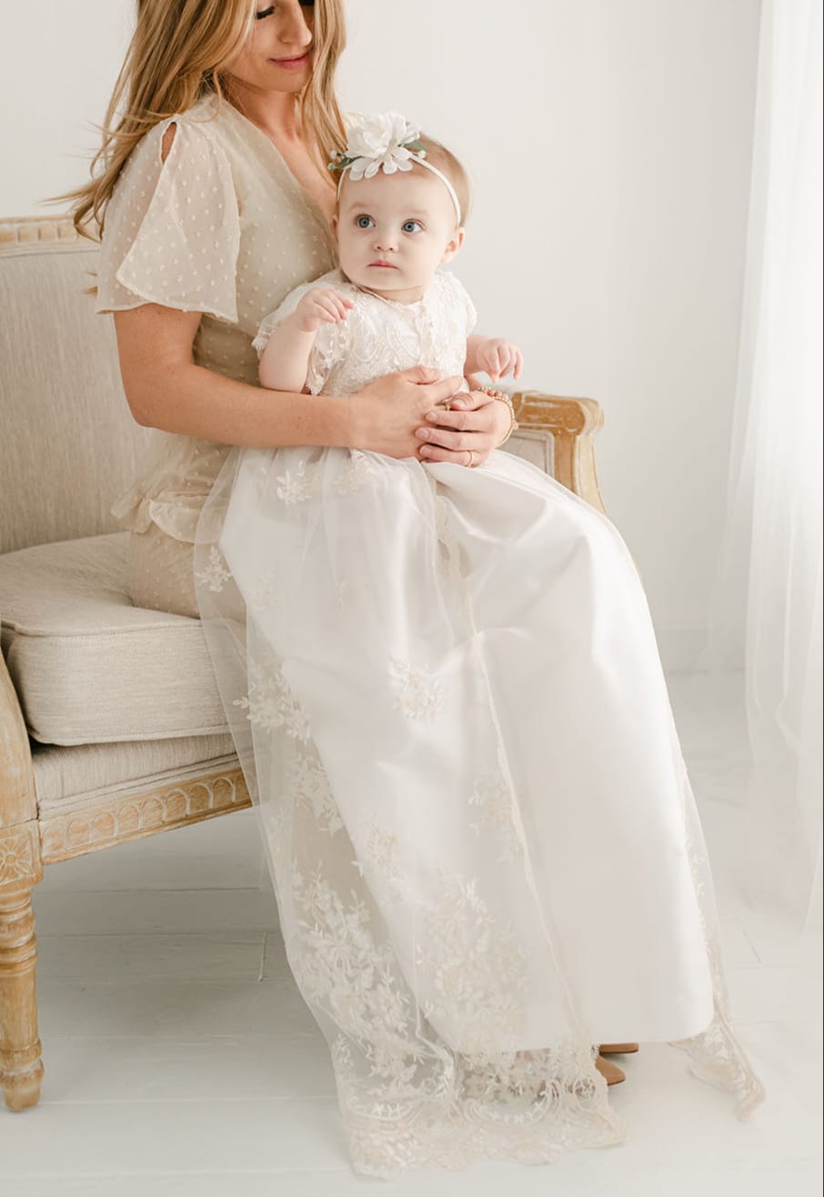 A woman sits on an elegant armchair holding a baby dressed in a white lace gown and headband for a christening, both looking towards the camera in a bright room