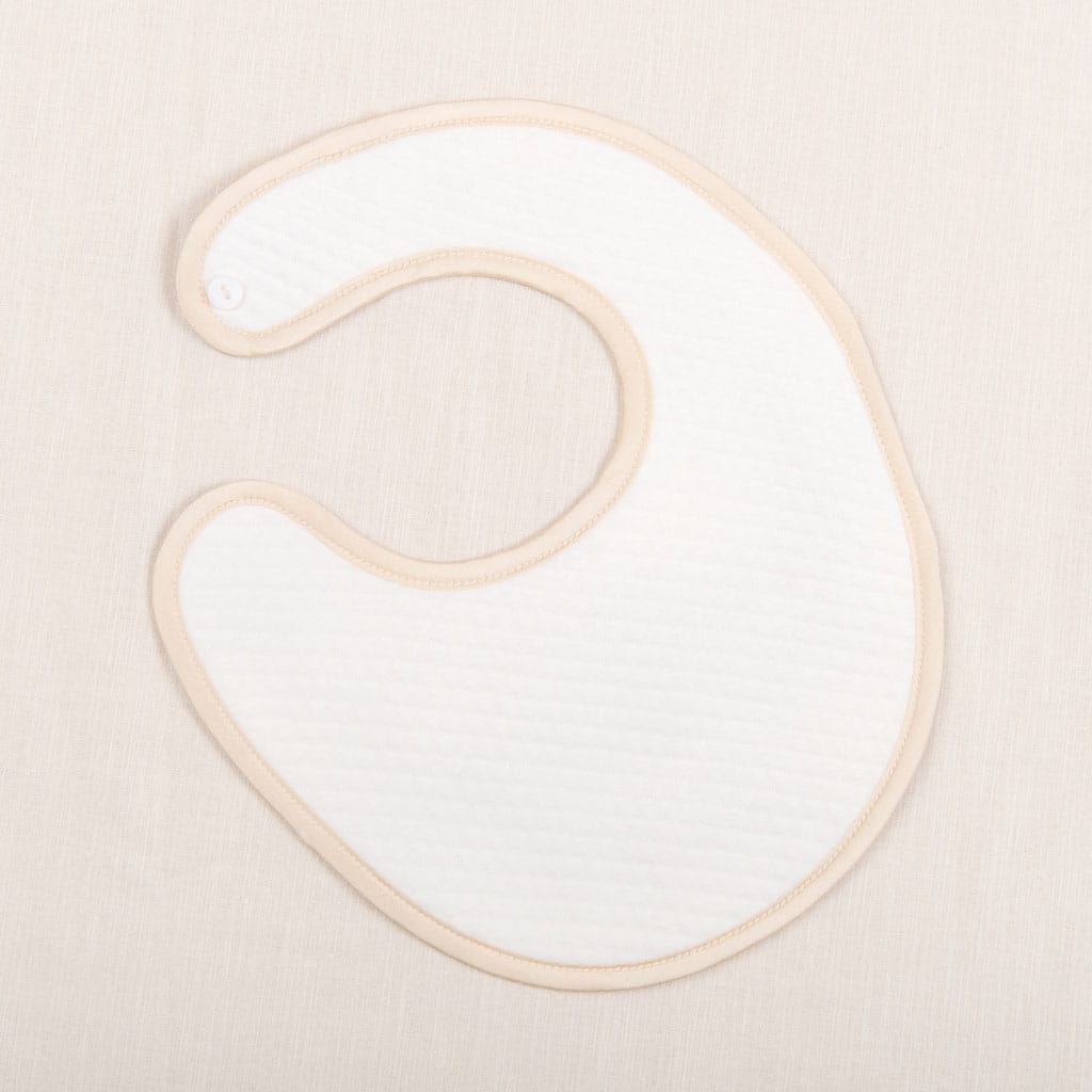 Flat lay photo of Liam Boys Bib. It is crafted with soft quilted cotton in ivory and is detailed with a silk champagne trim
