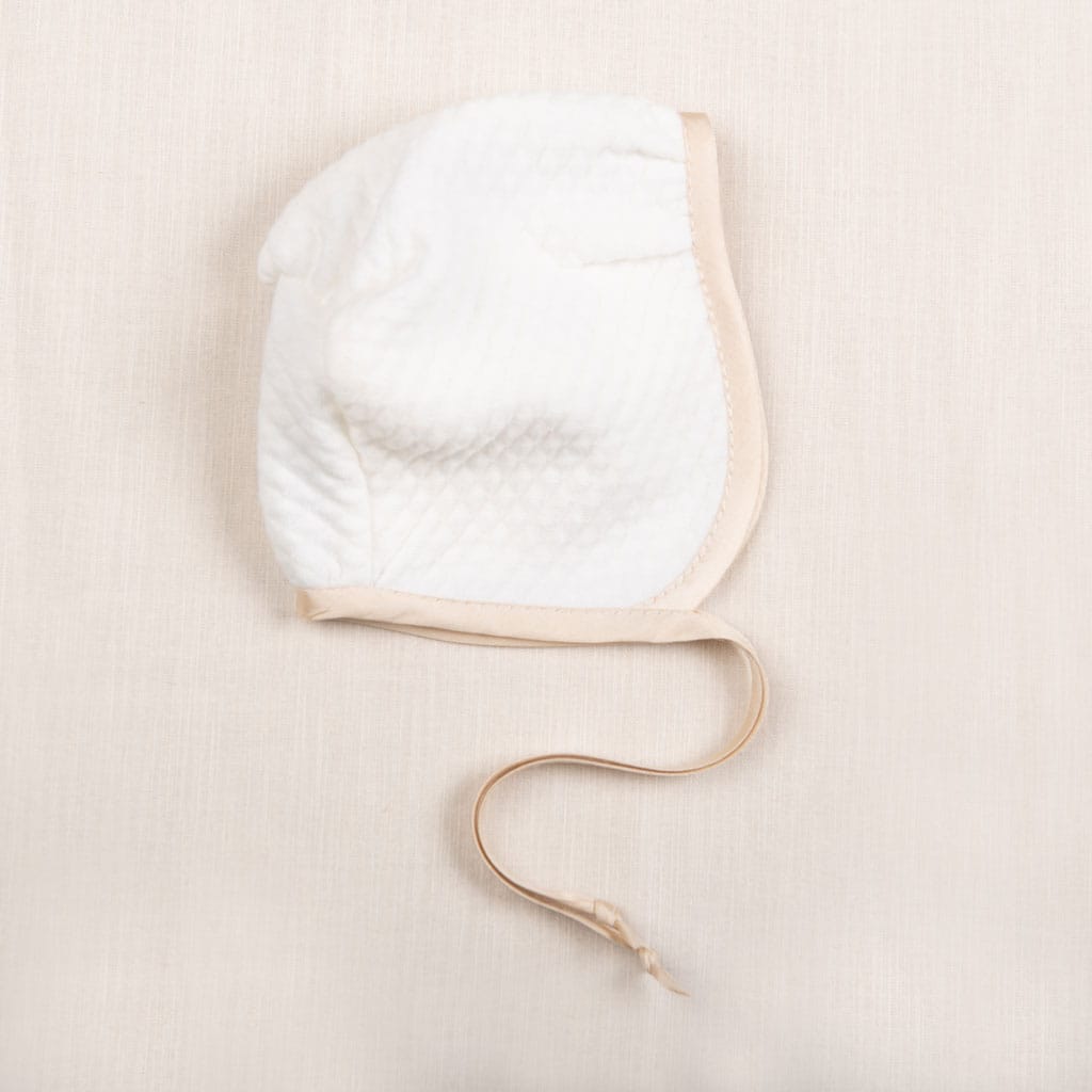 Flat lay photo of the Liam Quilted Newborn Bonnet made with soft quilted cotton in ivory and trimmed with a champagne colored silk