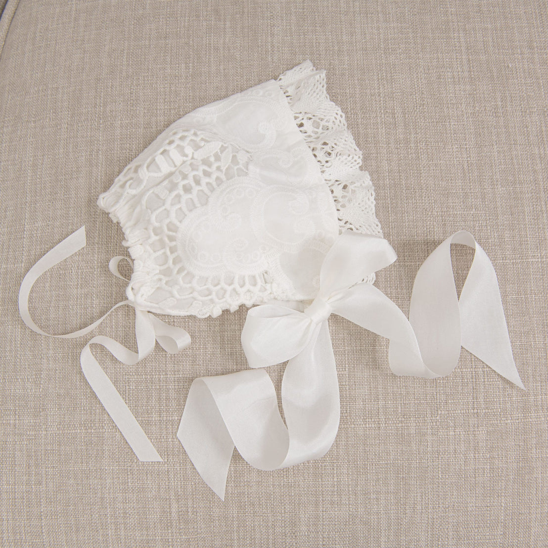 Flat lay of the Lily Bonnet on a beige fabric surface, highlighting the light ivory cotton lace details, edge lace, and ivory silk ribbons.