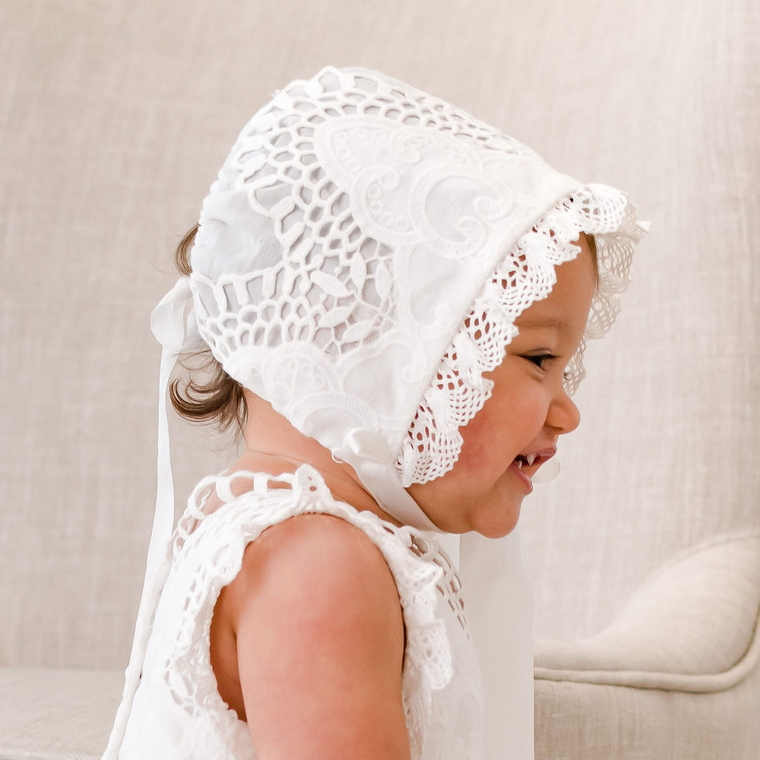 A baby girl wearing a Lily Bonnet and matching dress looks away from the camera with a big grin. The side profile of the girl showcases bonnet's ivory cotton lace details and edge lace.