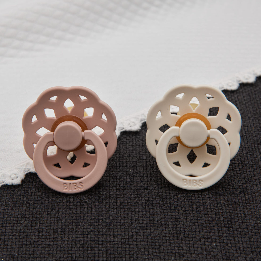Blush pink and ivory pacifiers from the Lola Pacifier Set displayed against a dark textured fabric, with a white quilted blanket in the background.