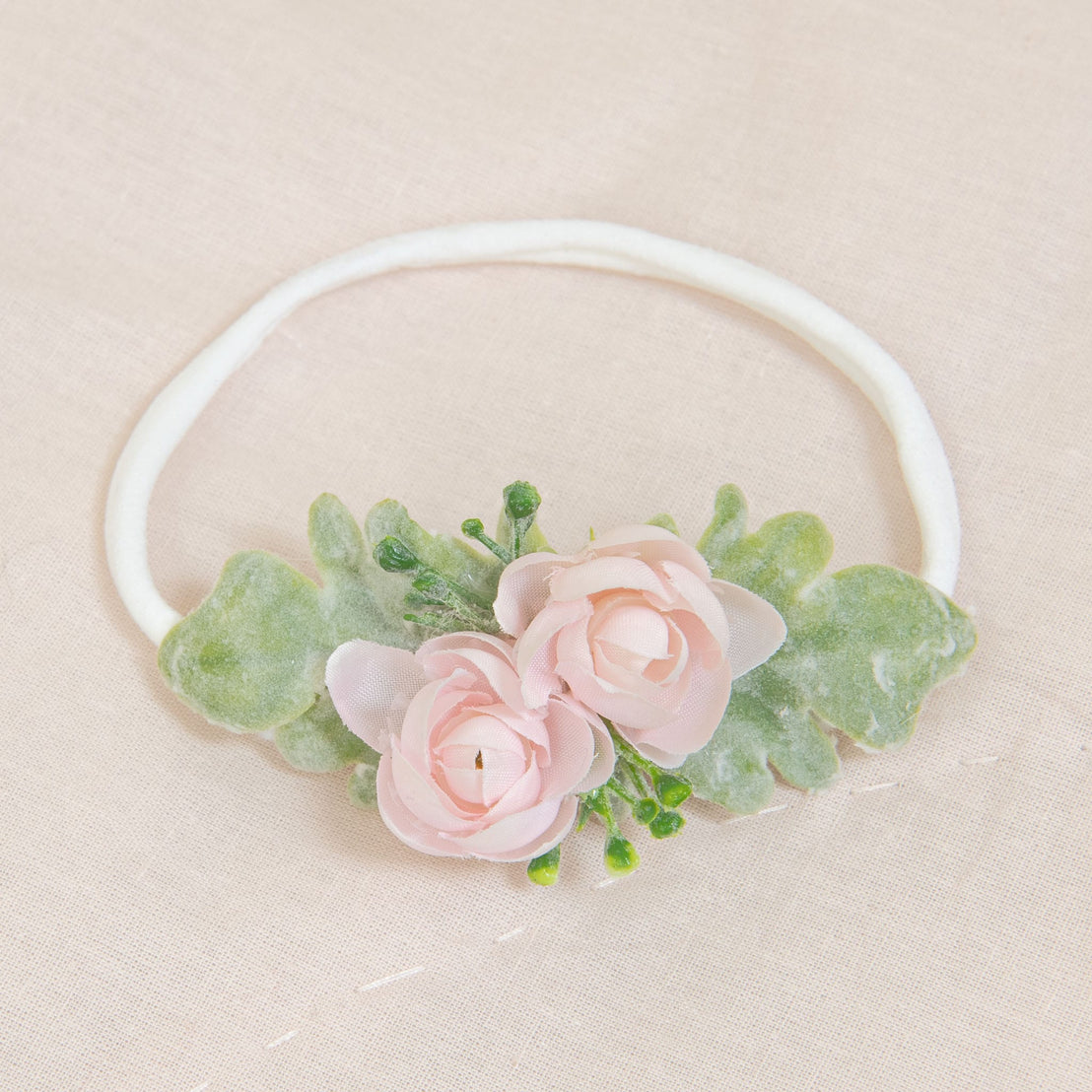 The Natalie Flower Headband showcases two pink roses and green leaves, beautifully arranged on a light beige fabric background, skillfully crafted from hand-stretched nylon.