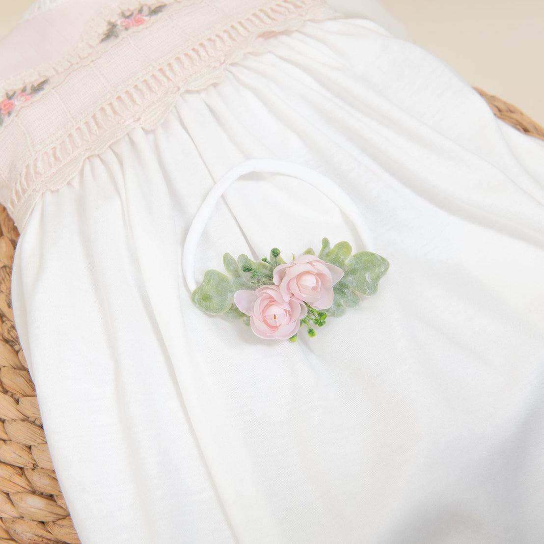 A white baby dress with pink embroidered flowers is laid on a wicker basket, complemented by the Natalie Flower Headband, which features handmade embellishments of pink roses and green leaves, creating a soft and delicate aesthetic.