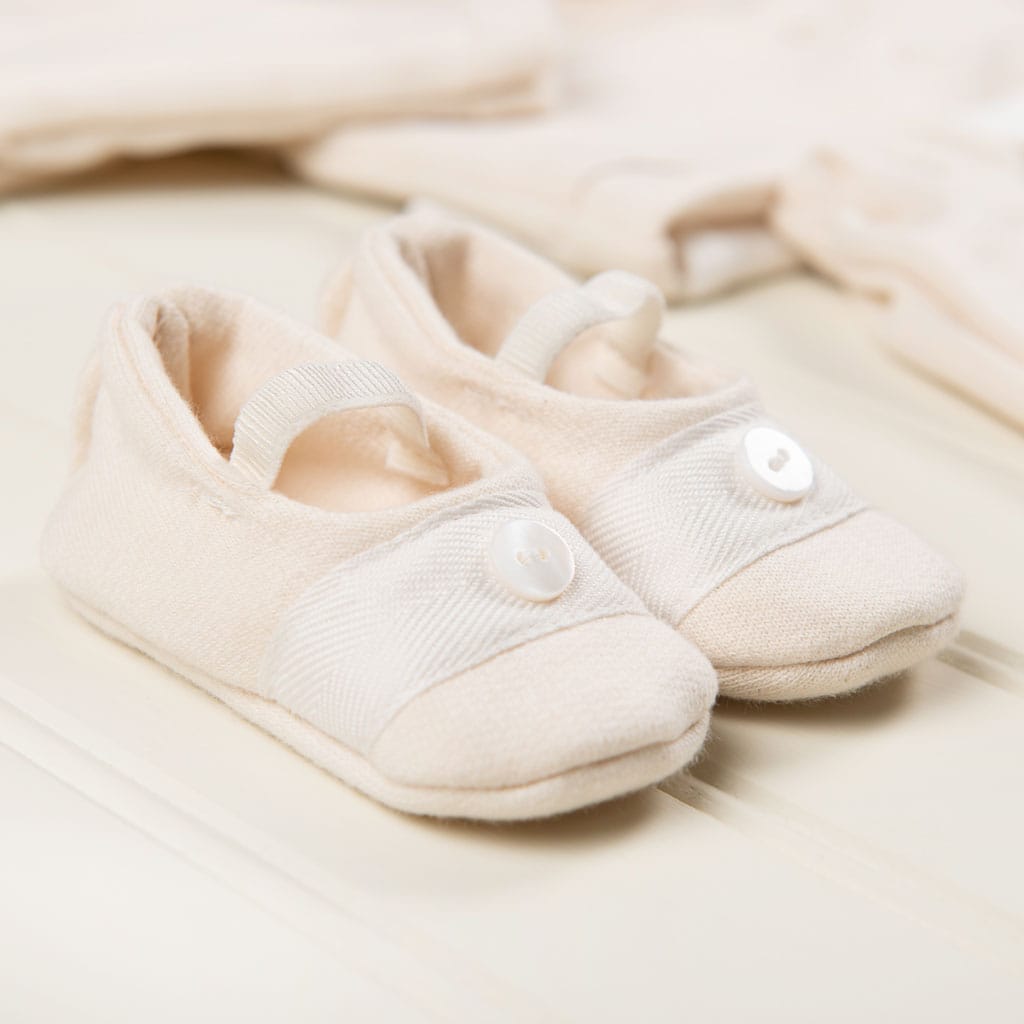 A close-up of Natural French Terry Booties, a creamy beige pair of baby shoes handcrafted in the USA. These feature soft French Terry fabric and a button detail on the front, resting on a light-colored wooden surface with blurred hints of other baby clothing items in the background.