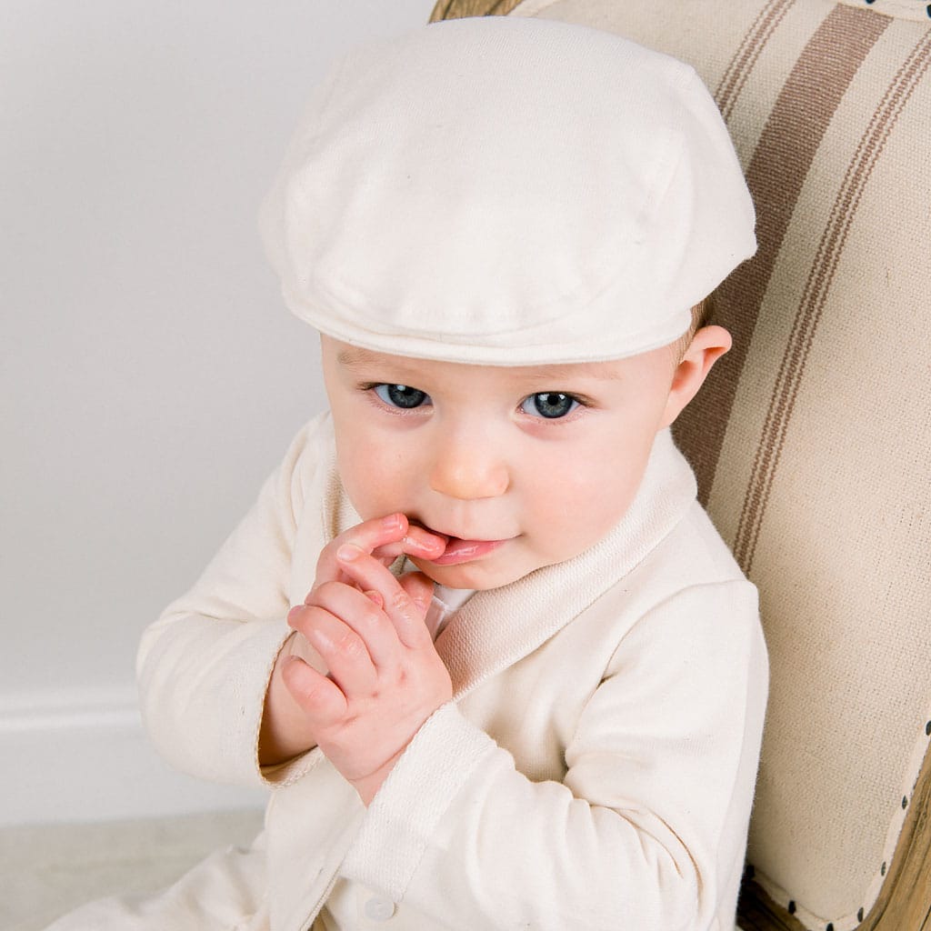 A baby wearing a cream-colored outfit made from soft French Terry Cotton, paired with a Natural French Terry Newsboy Cap, sits on a striped chair. The light-skinned, blue-eyed infant playfully touches their lips with their fingers while gazing curiously at the camera.