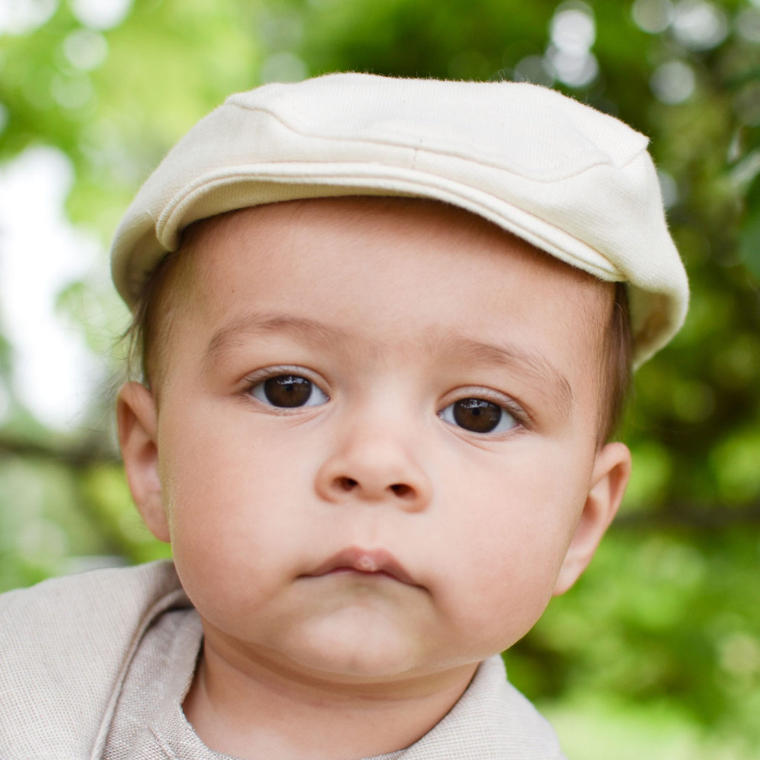 A baby, sporting a Natural French Terry Newsboy Cap, gazes directly at the camera. The cream-colored flat cap is crafted from French Terry Cotton, and the scene is set against a blurred backdrop of lush green foliage that evokes an outdoor vibe. This handmade ensemble from the USA features a neutral outfit with a soft elastic back for added comfort.