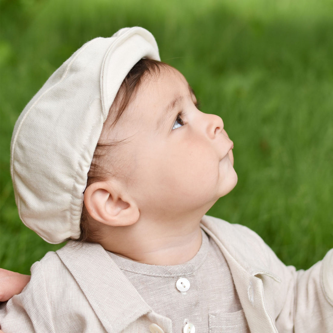 Dressed in a beige French Terry Cotton outfit, a baby wearing the Natural French Terry Newsboy Cap gazes upward against a blurred green background, with the soft elastic back offering both comfort and style.