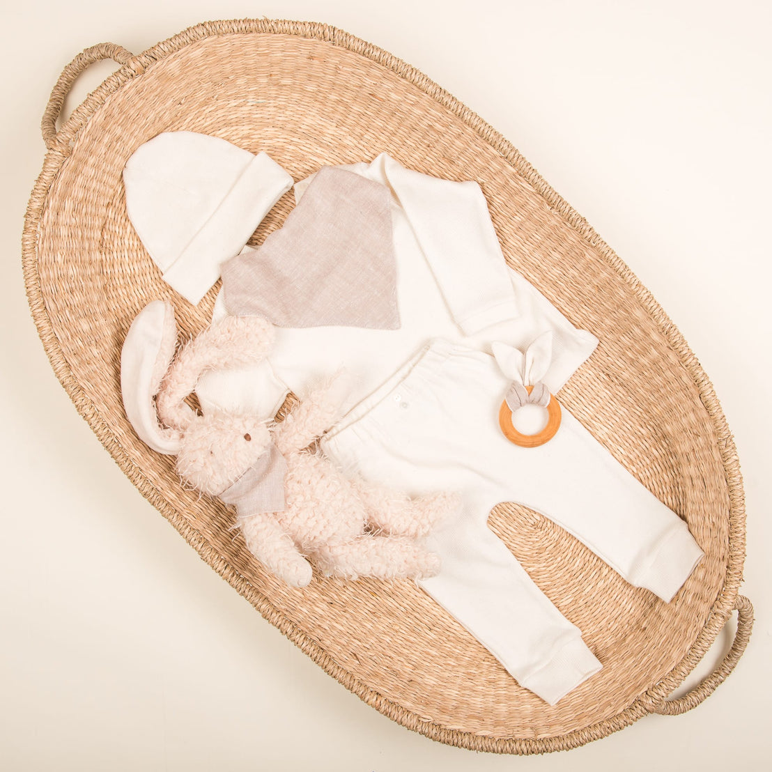 A wicker basket filled with a baby clothes set, featuring the Natural Ivory Ribbed Cotton Top & Leggings, includes a white hat and a beige bib. Completing the handmade in the USA outfit arrangement are a plush rabbit toy and wooden teether, all elegantly displayed on a neutral background.