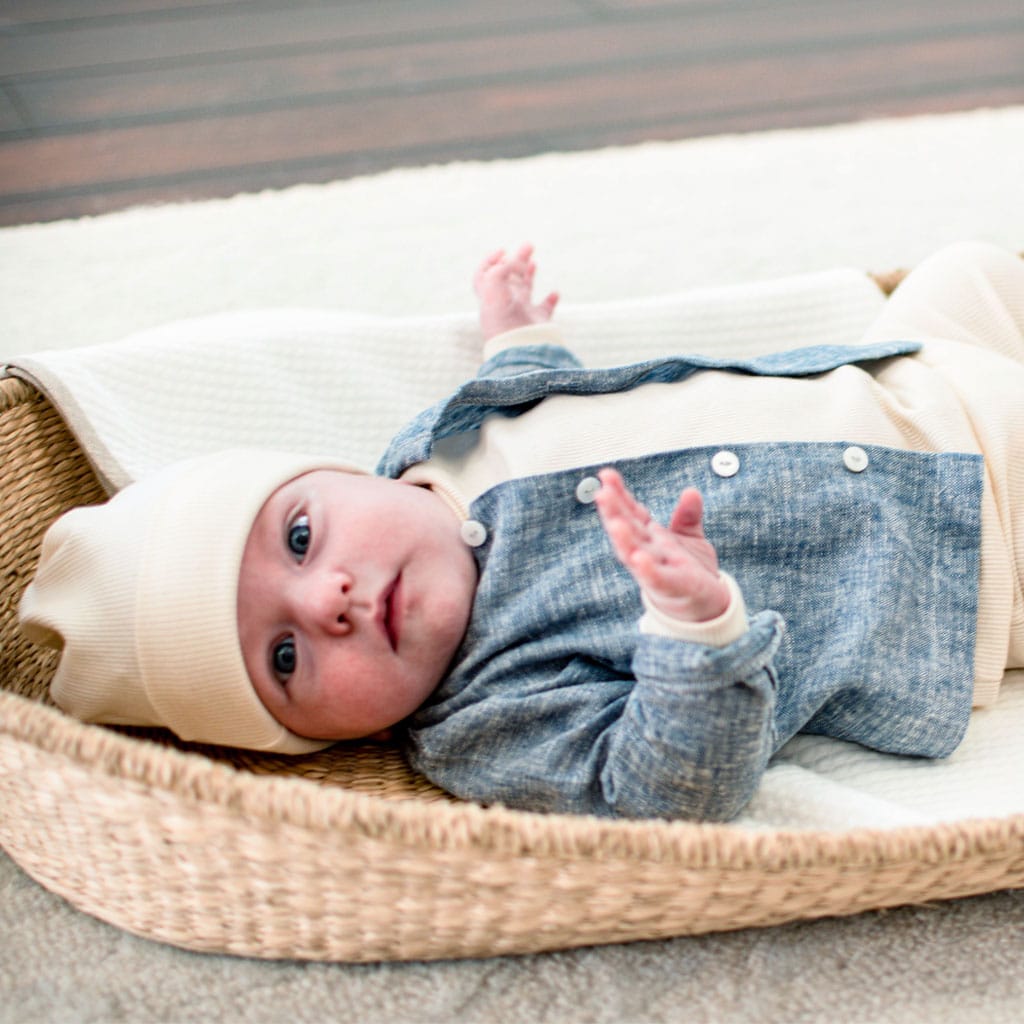 A baby dressed in a handmade in the USA Natural Ivory Ribbed Cotton Top and Leggings is lying in a wicker basket on a white blanket. The infant looks upward, with hands slightly raised. The setting appears cozy and warm, highlighting an outfit crafted with care.