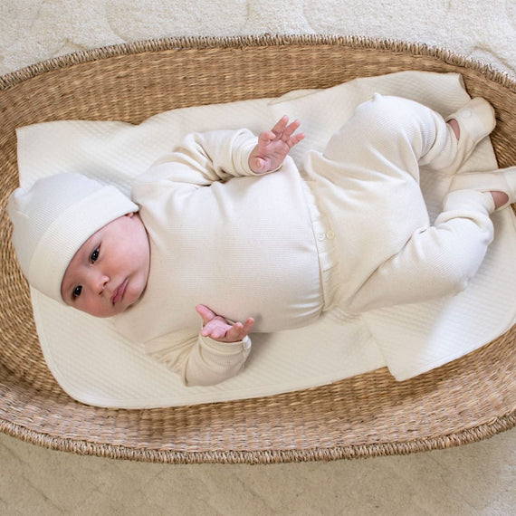 A baby dressed in the Natural Ivory Ribbed Cotton Top & Leggings and matching hat lies on a white blanket inside a woven basket. The baby gazes upward with their arms slightly raised, resting on a soft, light-colored surface that highlights the delicate craftsmanship handmade in the USA.