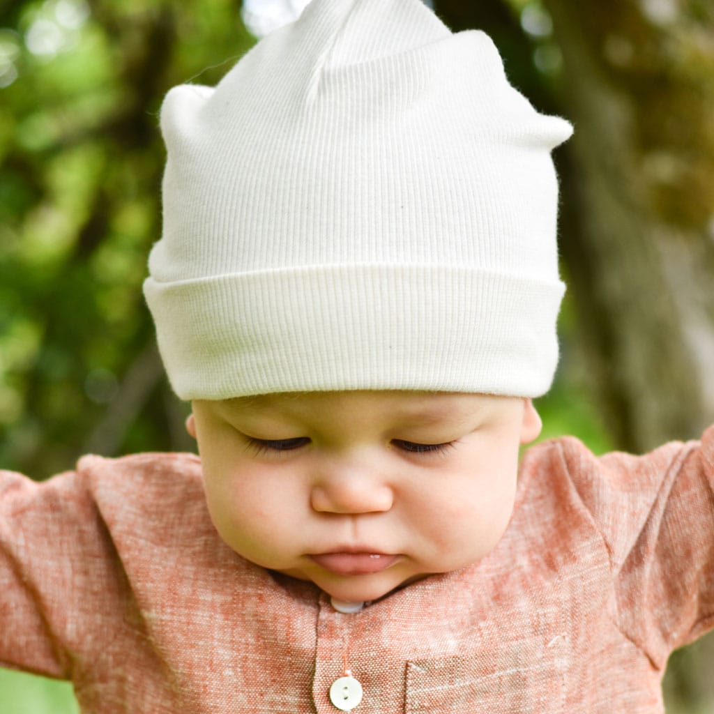 A baby sporting the Natural Ivory Ribbed Pima Beanie and a rust-colored shirt is looking down with their arms spread outward. The background features a blurred outdoor scene with green leaves and tree trunks, creating an enchanting setting for this handmade beanie.