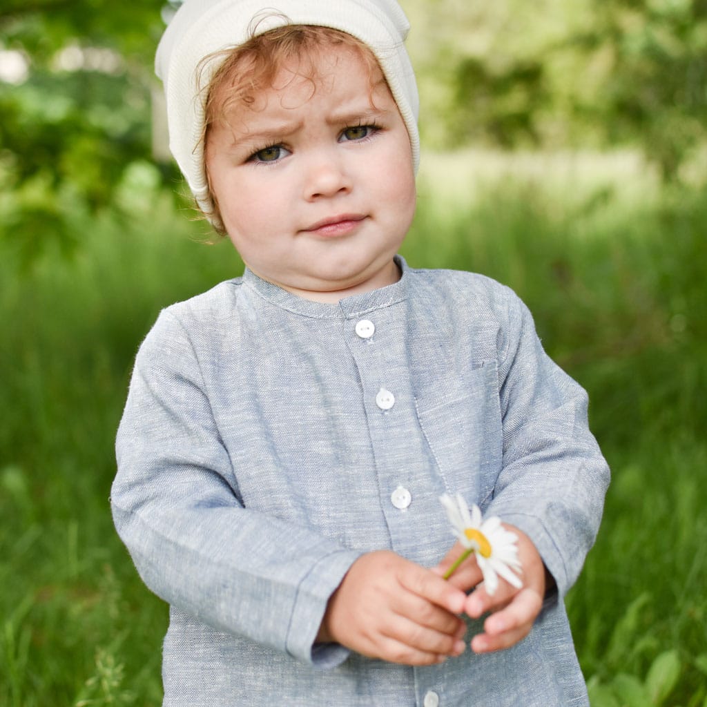 A toddler with curly hair and a Natural Ivory Ribbed Pima Beanie looks curiously at the camera while holding a daisy. They are wearing a light blue shirt made from ribbed Pima cotton and standing outdoors, with green foliage in the background.