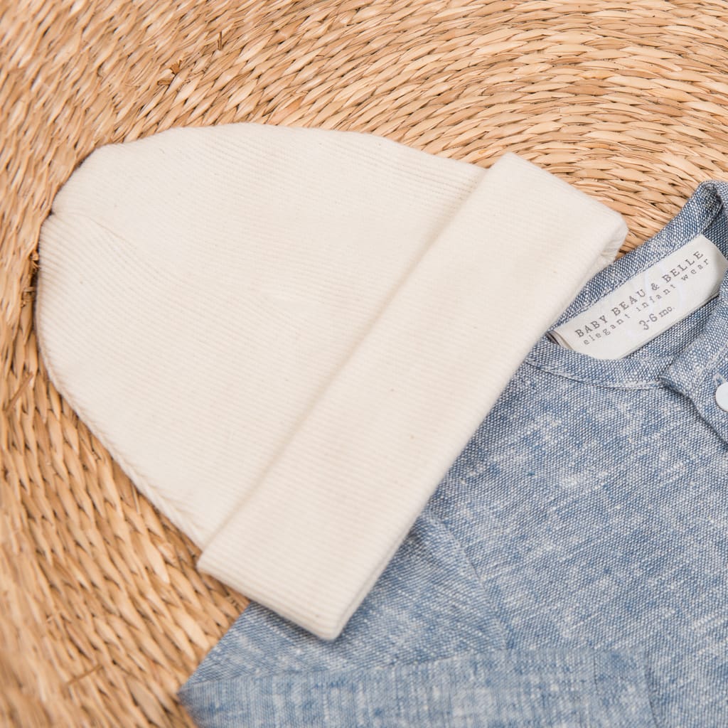 A Natural Ivory Ribbed Pima Beanie and a blue heathered baby onesie are neatly laid out on a woven rattan surface. The onesie, crafted from ribbed Pima cotton, has a label partially visible and features buttons on the front.