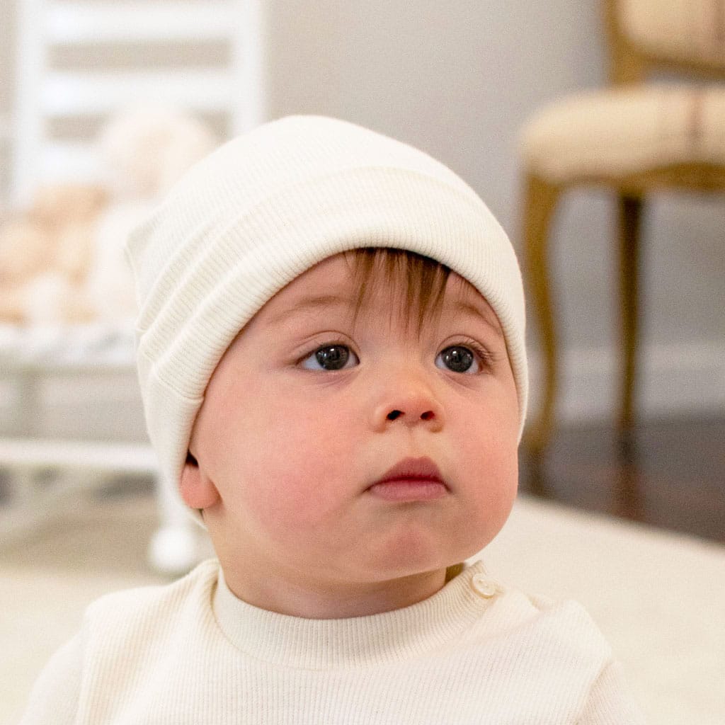 Wearing a Natural Ivory Ribbed Pima Beanie and a light-colored shirt, the baby gazes upward with a curious expression. The background showcases a blurred chair and crib, adding to the cozy indoor ambiance.