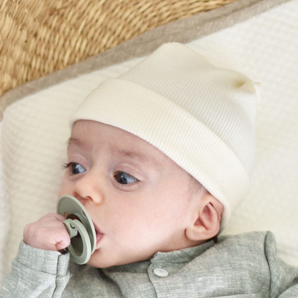 A baby wearing a Natural Ivory Ribbed Pima Beanie and a green outfit holds a green pacifier in their mouth. The baby is lying on a soft, textured surface and looking off to the side.