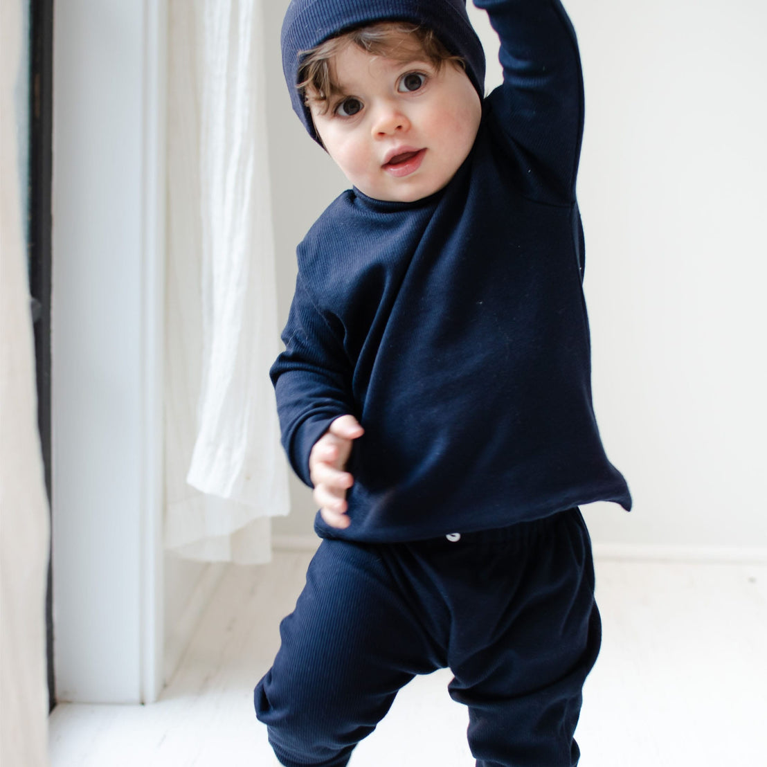 Baby boy standing and wearing the navy ribbed cotton top and leggings (and Navy Ribbed Pima Beanie). The top and leggings are made out of 100% ribbed textured cotton with button detail on leggings. 
