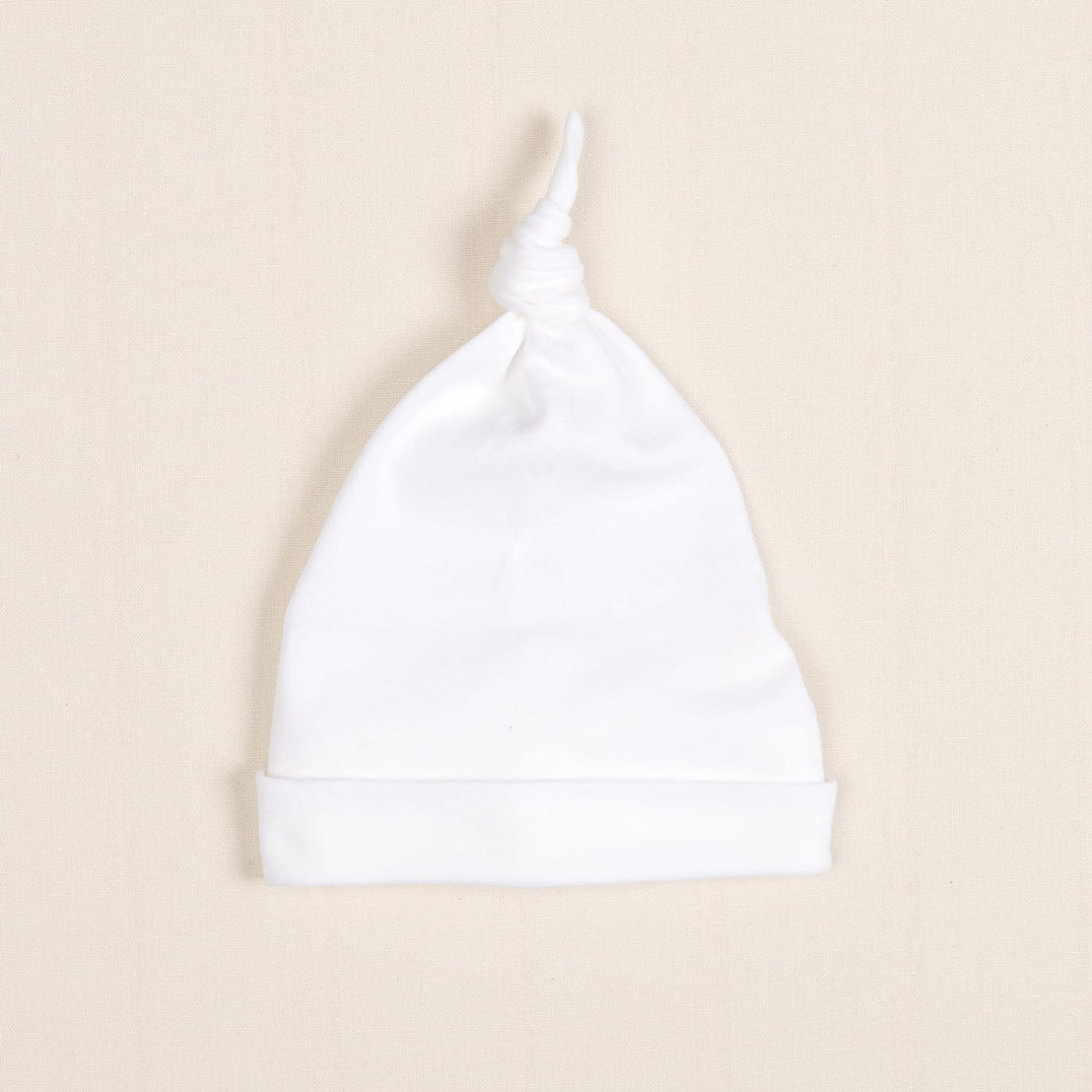 The Newborn Knotted Cap, also known as the Liam Cap, is displayed flat on a beige background, making it an ideal choice for a soft and stylish newborn outfit.