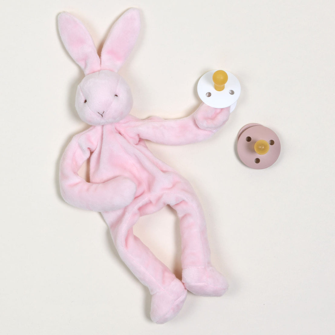 A Pink Silly Bunny Buddy & Pacifier, made to be safe for babies, is situated on a light surface, clutching a white pacifier. Beside it lies a brown pacifier. The bunny with the pacifier flaunts floppy ears and minimalist embroidered details.